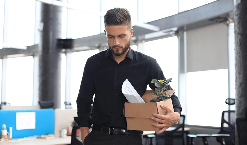 A man wearing a black shirt is walking away from his office and carrying a box with paperwork and a plant in it. This man has received a dismissal from his workplace.
