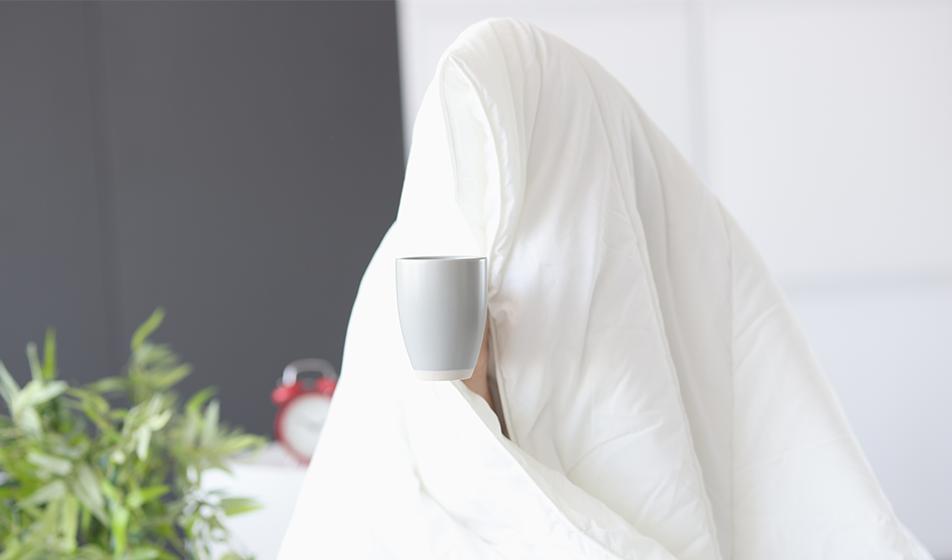 Someone is enveloped in a white duvet that obscures their face. A cup that they're holding can be seen. 