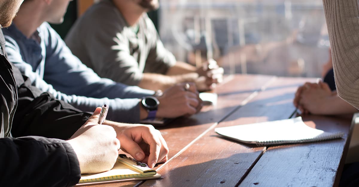 Four colleagues are sat around a wooden table together. They are having a meeting and writing things down in their notebooks.