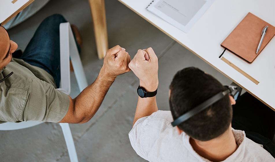 Two male colleagues are shown from above, both sat at their office desks. One has a headset on. They're shown to be fist-bumping, showing friendship, trust, or appreciation. 