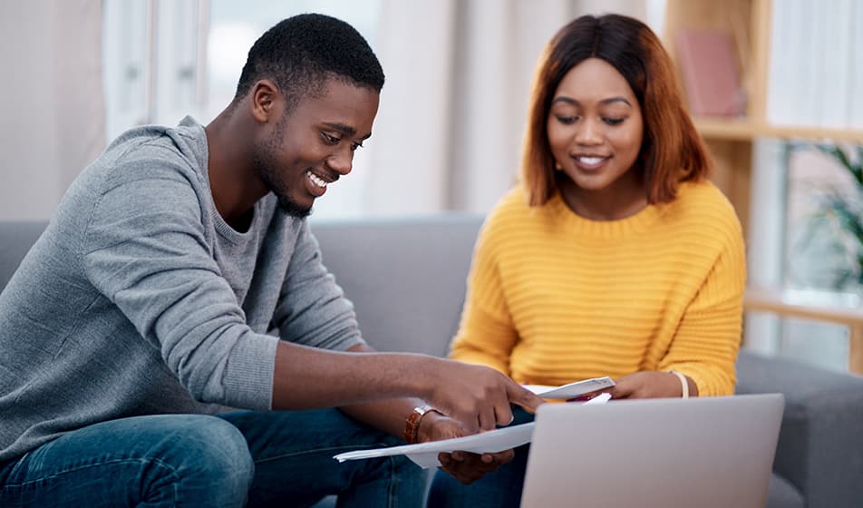 A man and a woman sit on sofa with a laptop on a coffee table in front of them. They are both reviewing some paperwork, smiling.