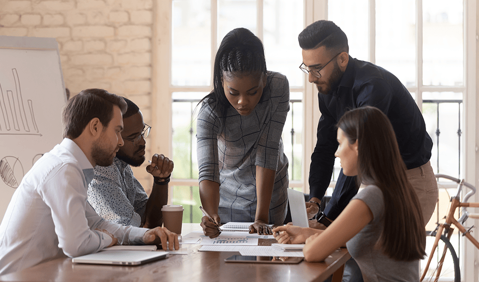 Five colleagues are working together around a table. Two of the colleagues are stood up and the rest of them are sat down. They are all looking at a piece of paper with a graph on. Behind them is a stand holding an A3 sized piece of paper on. 
