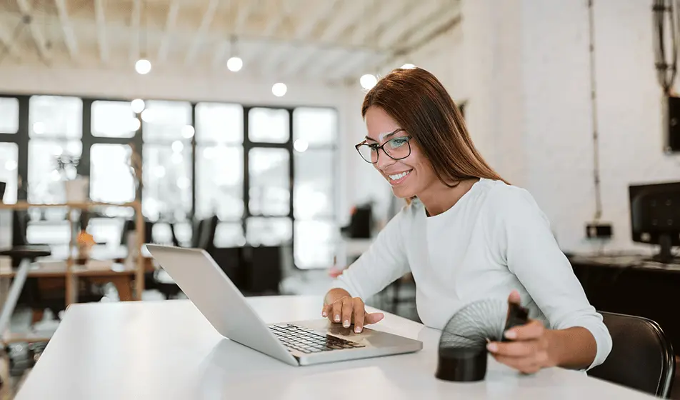 A woman with glasses is in an office, sitting with her laptop at a white desk. She is looking at her laptop and is playing with a black slinky on the table.