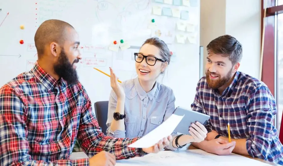 Three colleagues are sat working at a desk together. Two of them are men wearing flannel shirts and one is a woman wearing a blue shirt and glasses. They are laughing in the office.