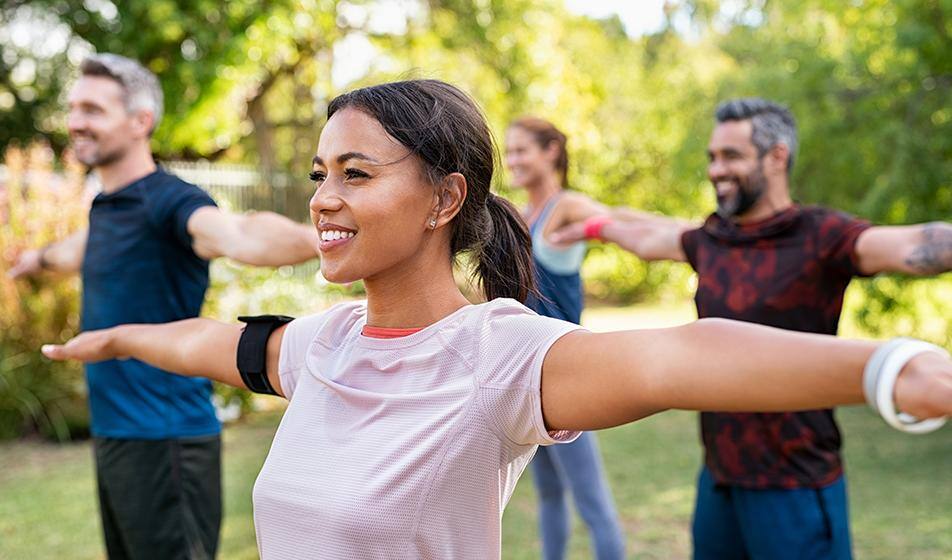 A group of men and women are standing in a yoga pose, arms outstretched in a 'wingspan' pose. They are outside amongst trees & are all smiling. 
