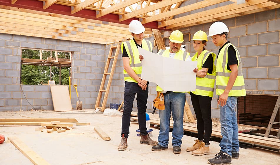Four people are stood in a construction site and are wearing hi-vis jackets with hard hats. They are looking at a large piece of paper that has the plans for the building work on it.