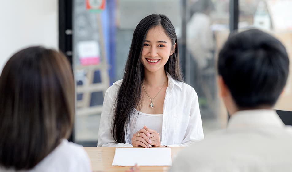 A smiling woman sits across from a man and a woman who have their backs to the camera, in an office interview environment. 