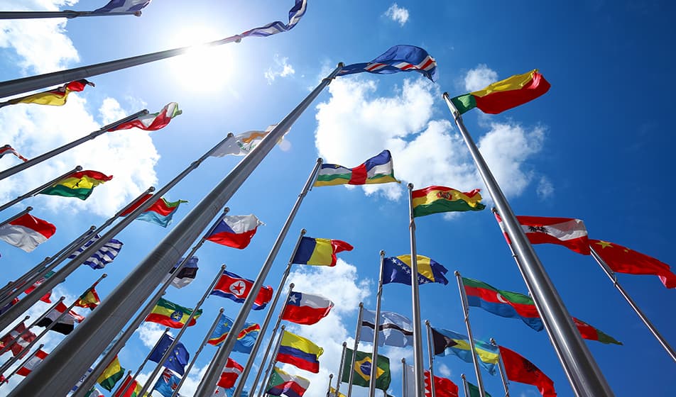 A group of international flags from all around the world are shown from below, flagpoles reaching up to the blue sky, containing a few white clouds. 