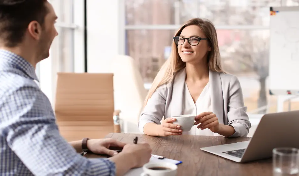 Two colleagues are sat at a table together having a meeting. The female colleague is sat next to her laptop and is smiling at her male colleague who is smiling back and writing notes.