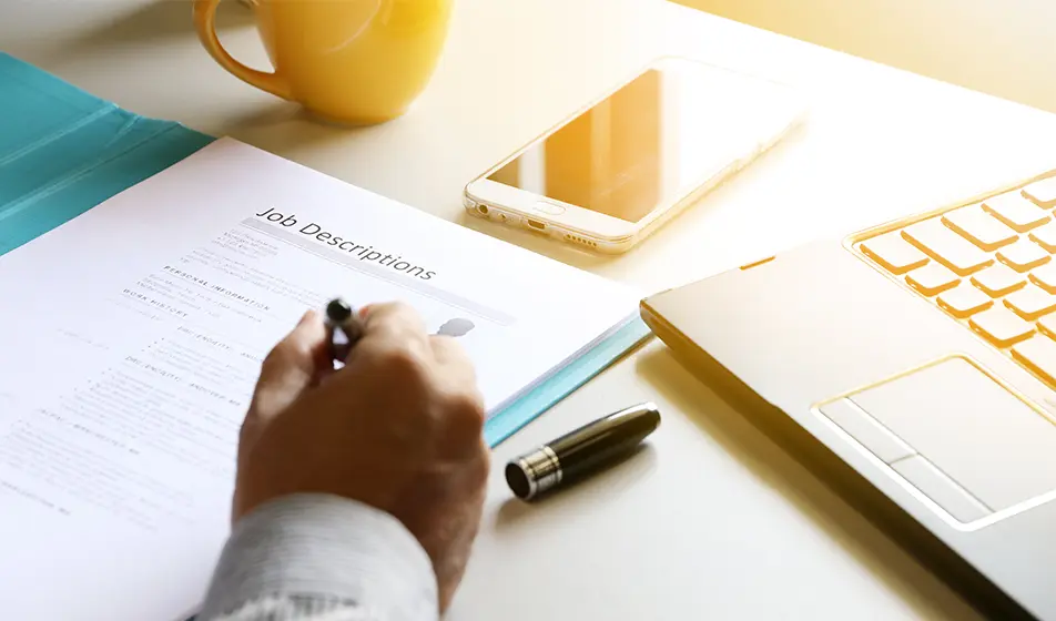 A document titled 'job description' is shown on a wooden table, with a laptop, a smartphone and a yellow mug nearby. A man's hand can be seen holding a pen over the document.