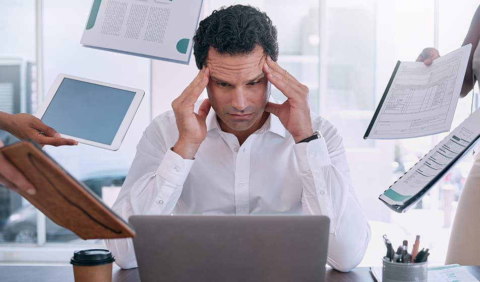 A man sits in front of his laptop, hands either side of his temples, looking stressed. Either side of him, people are presenting him with documents and tablets, displaying an overwhelming image. 