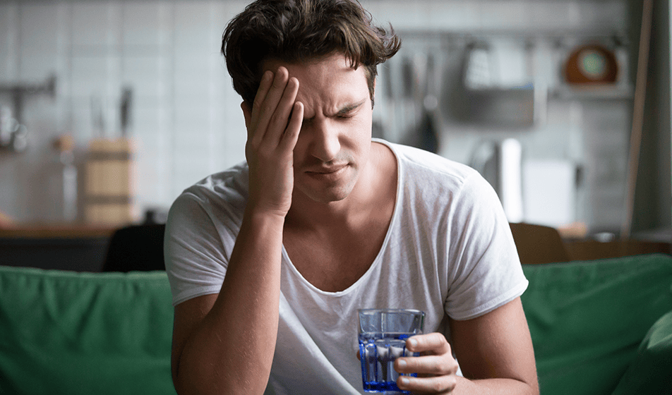 A man is suffering from long-term sickness. He is sat on a green sofa with his hand on his head and a glass of water in the other hand.
