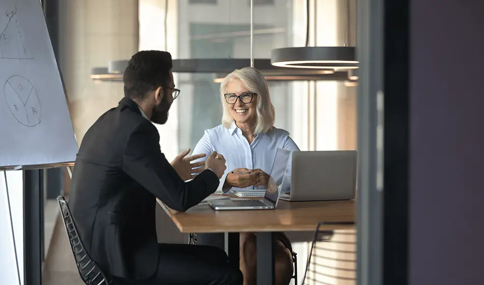 A man and a woman are sitting at a wooden desk with laptops and paperwork in front of them. They are having a meeting in one of their office rooms, smiling and chatting to each other. 