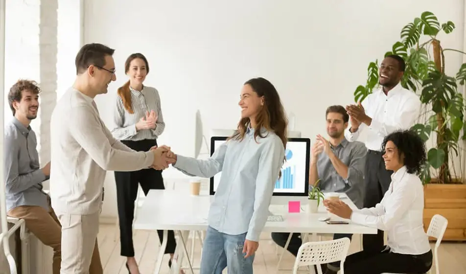 A woman is shaking the hand of her male colleague and smiling. Surrounding them are more colleagues who are clapping.