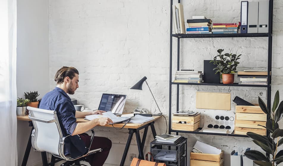 A man is sat working at his wooden desk in a contemporary office. He is surrounded by shelves that have files and plants on. 