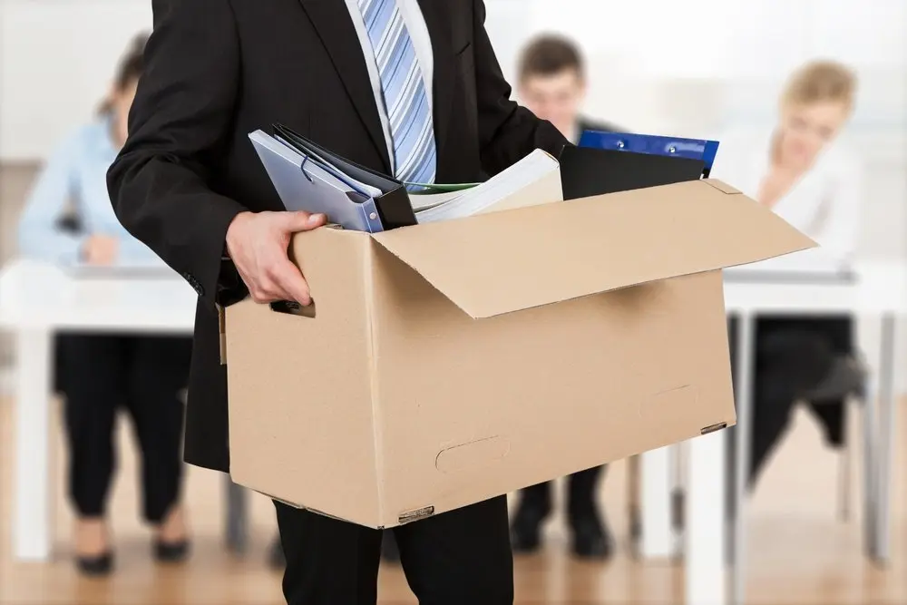 A man wearing a black jacket and a blue striped tie is holding a cardboard box and walking out of the office. Within his box are his belongings from work which include all of his folders and books.
