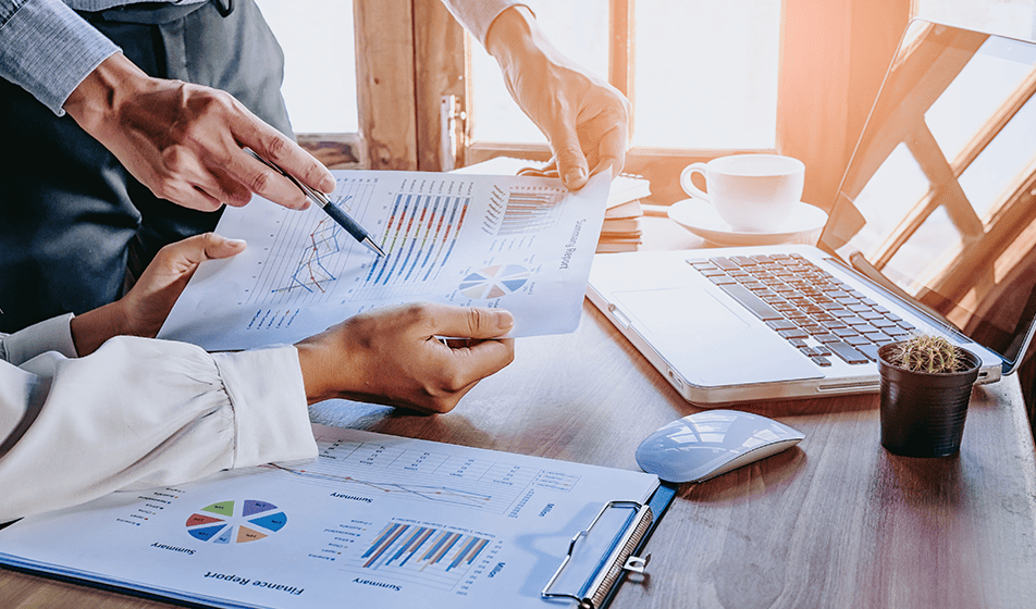 Two people are sat at a wooden table and looking at different graphs together. One person is pointing at a bar graph with a pen.