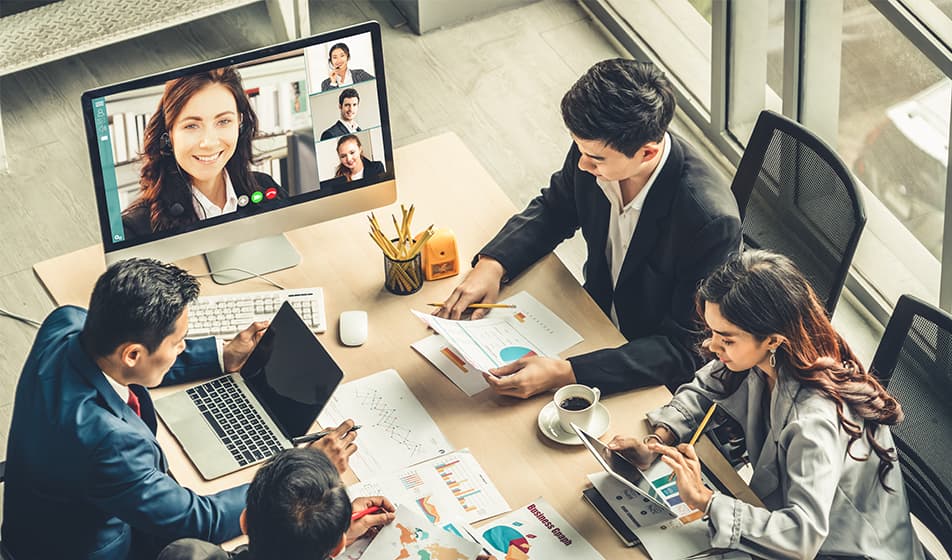 A group of employees sit around a table with laptops and papers in front of them. At the end of the table, employees join the meeting virtually via a screen.