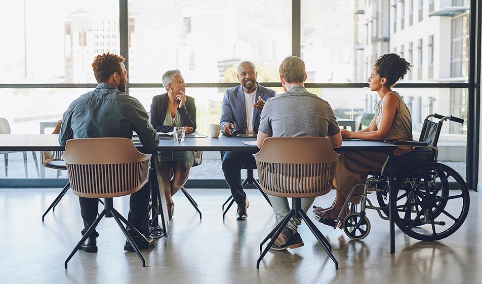 A group of diverse employees sit around a table in an office. An older lady is amongst them, as well as a lady in a wheelchair. They seem to be having a positive, calm discussion.