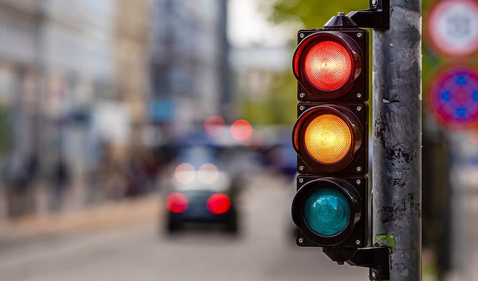 A traffic light is shown, showing red and amber lights illuminated. In the background and out of focus, a city scene can be seen, showing a car. 