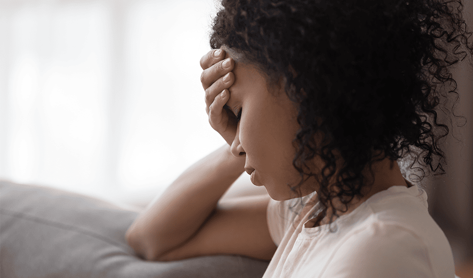 A woman wearing a white t-shirt is sat on a grey sofa and holding one hand to her head.