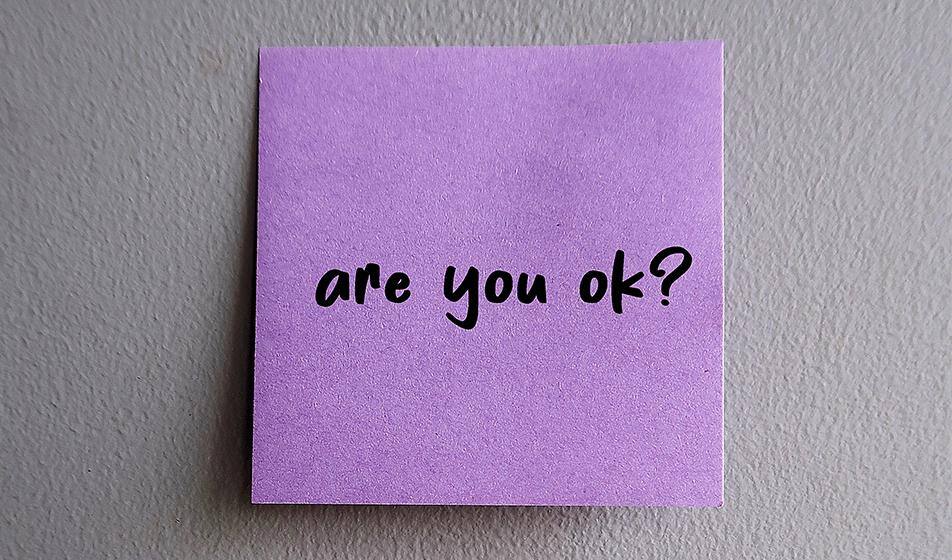 A lilac purple post-it note is stuck to a white wall. In black text, it reads, 'are you ok?'