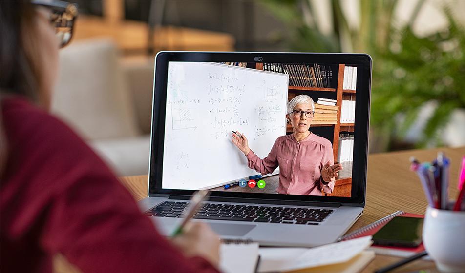 A woman sits at a desk in front of a laptop, where a woman presents training on screen. The woman at the desk is taking notes.