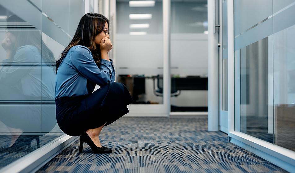 A woman is crouched down with her back against a glass office wall, with her chin in her hands. Her eyes are closed in an exasperated expression. This & her body language seem to say "I can't do this any more."
