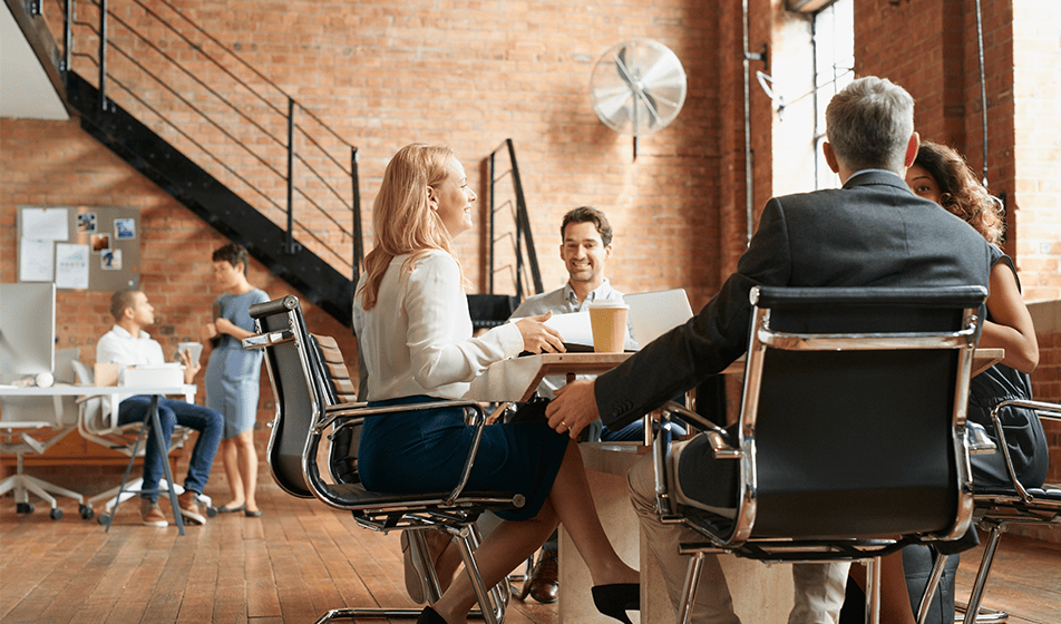 Several colleagues are in a modern, brick-walled office space. Four colleagues are sat in black chairs at a desk. They are talking within their group and smiling at each other. At the back of the office are two more colleagues. One is sat at a desk and the other is stood talking next to them.