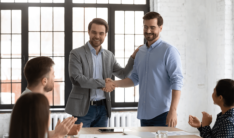 Four colleagues are in an office together. Two colleagues are sat at a table together looking up at the two colleagues who are stood up. One of the men stood up is shaking the others hand and introducing him to the group. They are onboarding a new employee.