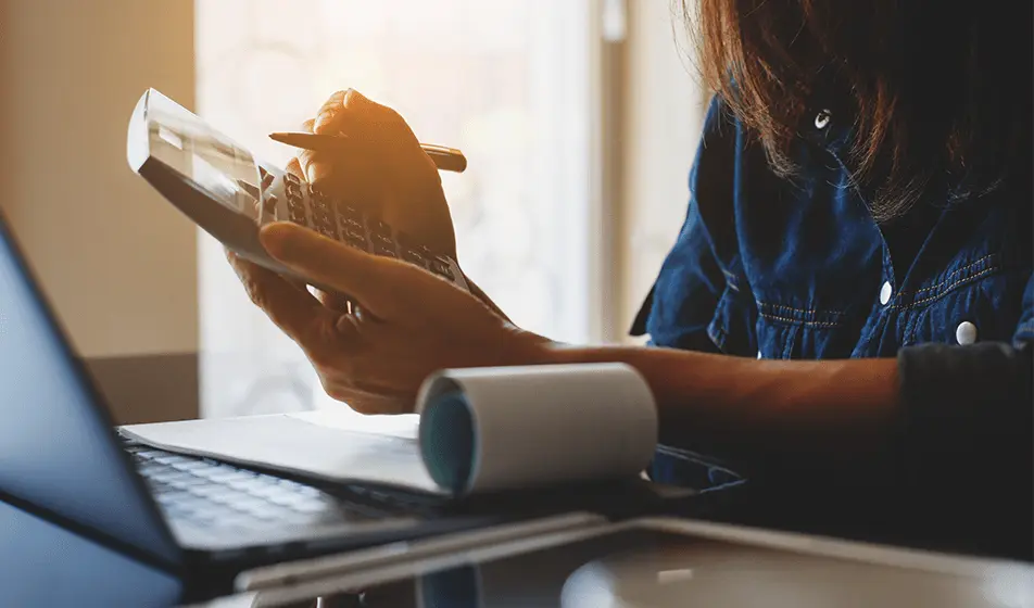 A woman's hair, arms and hands can be seen. She is holding a calculator and a pen in her hands. She has a small roll of paper on top of a laptop which sits in front of her.