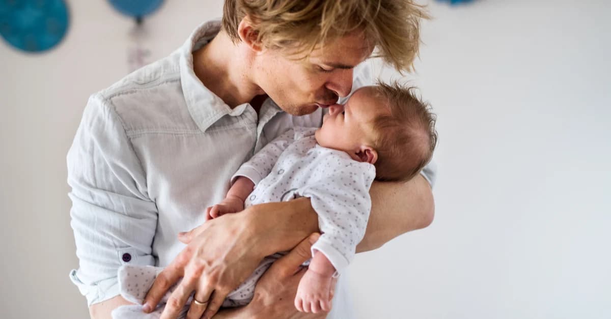 A father on paternity leave is holding his baby in his arms and kissing their forehead. Behind them is a white wall with blue balloons painted on it.