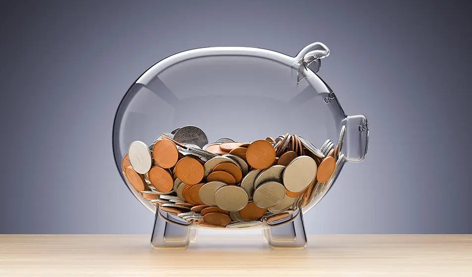 A transparent glass piggy bank is shown, with silver and bronze coins inside it up to around halfway. The background is grey and the piggy stands on a wooden table.