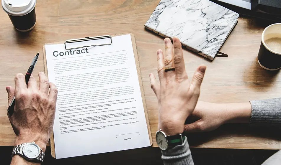 An image of contract on a clipboard. The contract is on a desk and two people are discussing the contents of it.