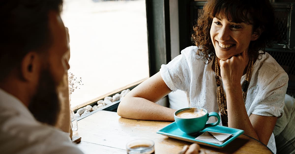 A man and a woman sat across from each other at a small circular table. They both have cups of coffee and are smiling.