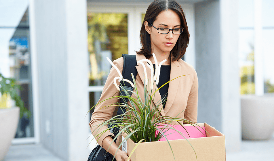 A downcast-looking woman walks from a building carrying a box containing office supplies and a plant. 