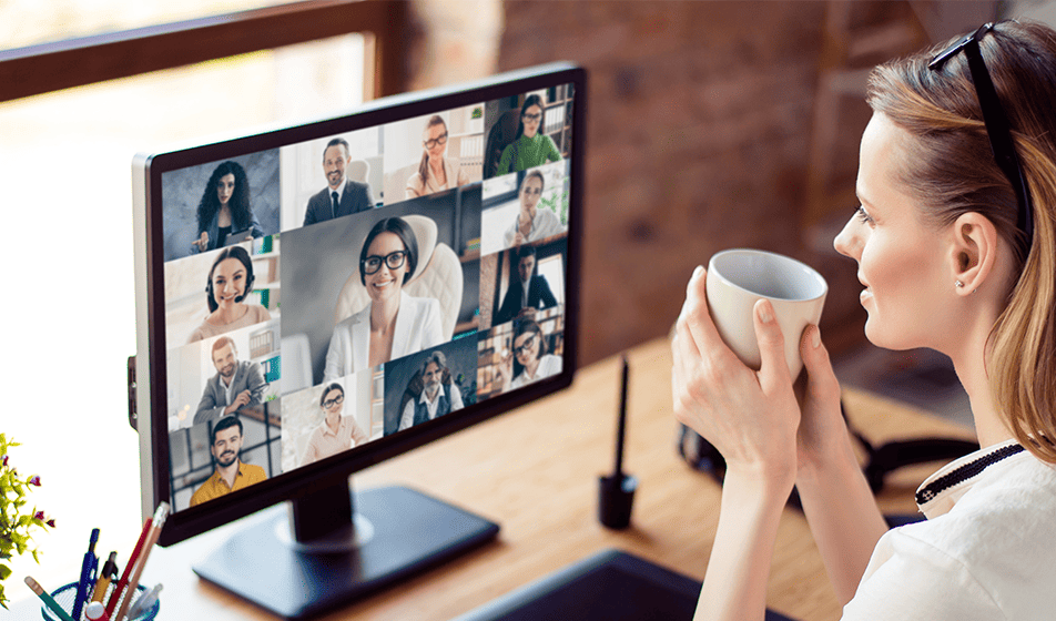 A woman works from home, looking at her colleagues on her screen via a virtual meeting. She holds a cup of coffee and seems relaxed. 