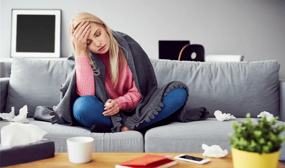 A woman is feeling unwell with the flu and cannot return to work yet. She is sat on a grey sofa, wrapped in a grey blanket with a hand on her forehead. She is surrounded by tissues.