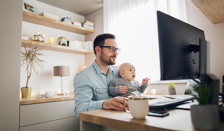 A man works from home in front of a computer monitor. He has a baby on his lap. 