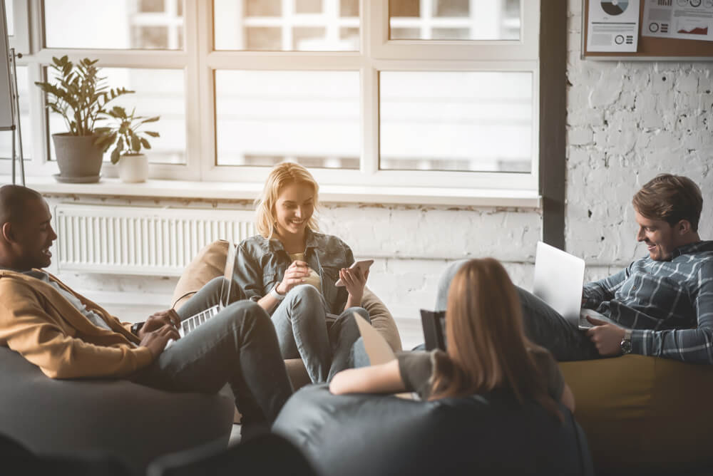 Four colleagues are sat on bean bags in a white-walled office. They are sat right next to a window which has two plants next to it.