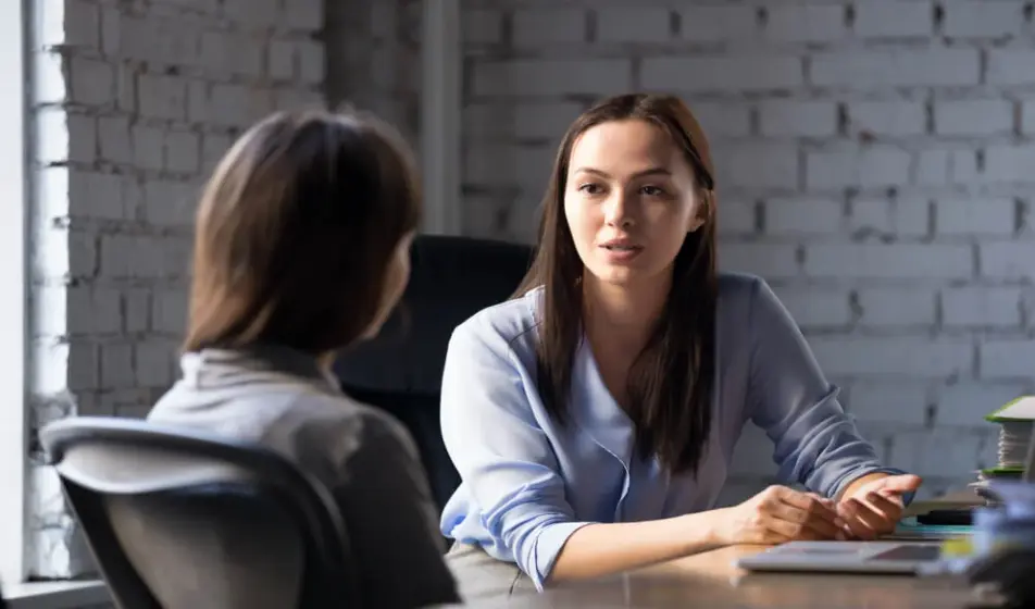 Two female colleagues are sat together at a wooden desk with paperwork piled in front of them. They are having a one to one meeting.