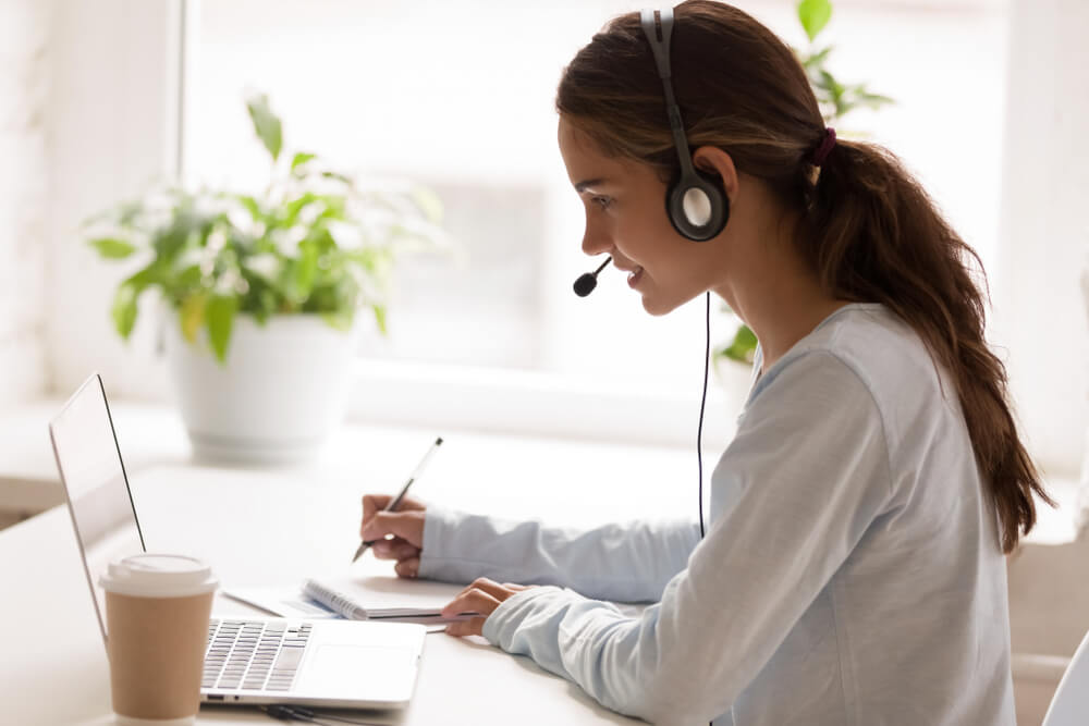 A woman is working from home. She has headphones on and is having a work call on her laptop which is in front of her. She has a takeaway coffee next to her and is writing notes down in her notebook.