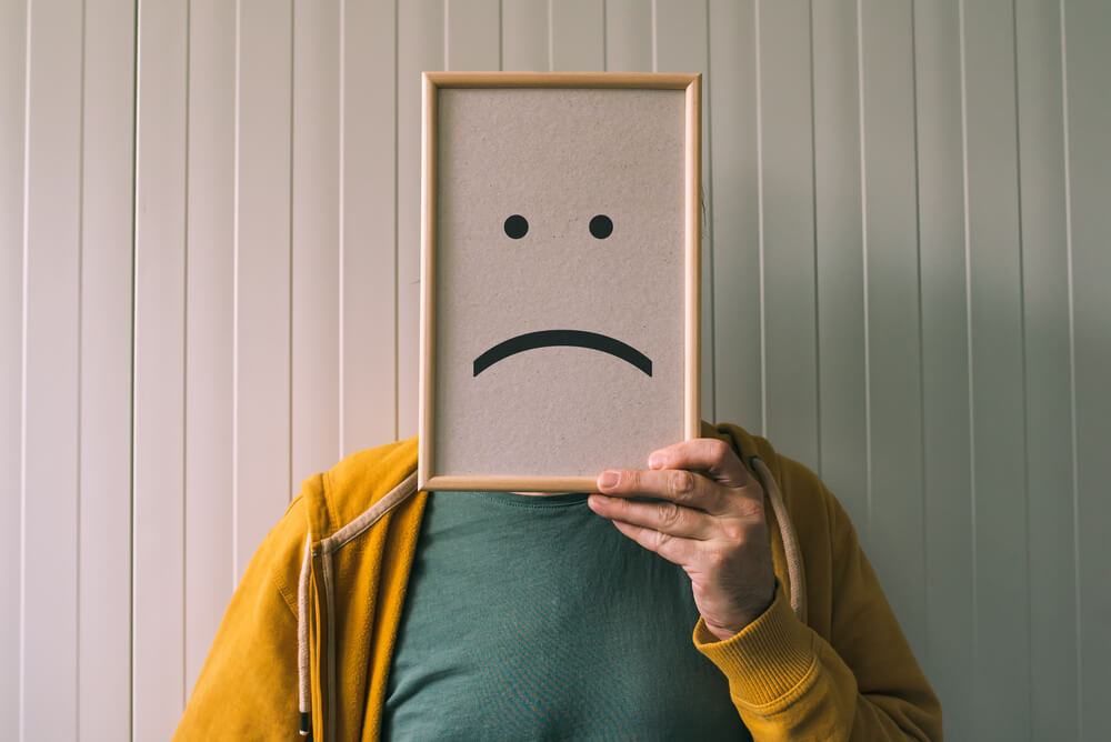A man in a yellow jacket and green shirt is standing against a white panelled wall. He is holding up a picture in front of his face that has a sad face on.