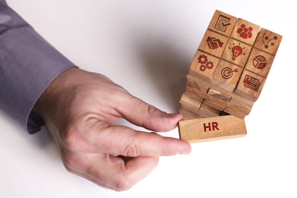 A man in a suit is playing jenga. The jenga pieces all have different symbols on them related to business such as a lightbulb. The man is pulling out a centre piece from the centre which has "HR" written on it.