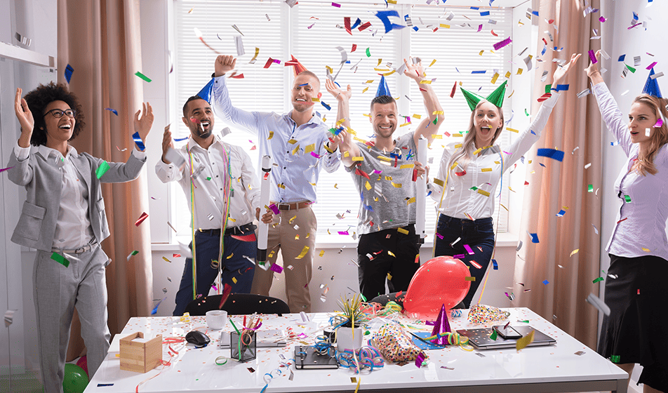 A group of employees stand to one size of an office desk that's been decorated & covered in balloons. They're smiling, wearing party hats & there's confetti in the air. 