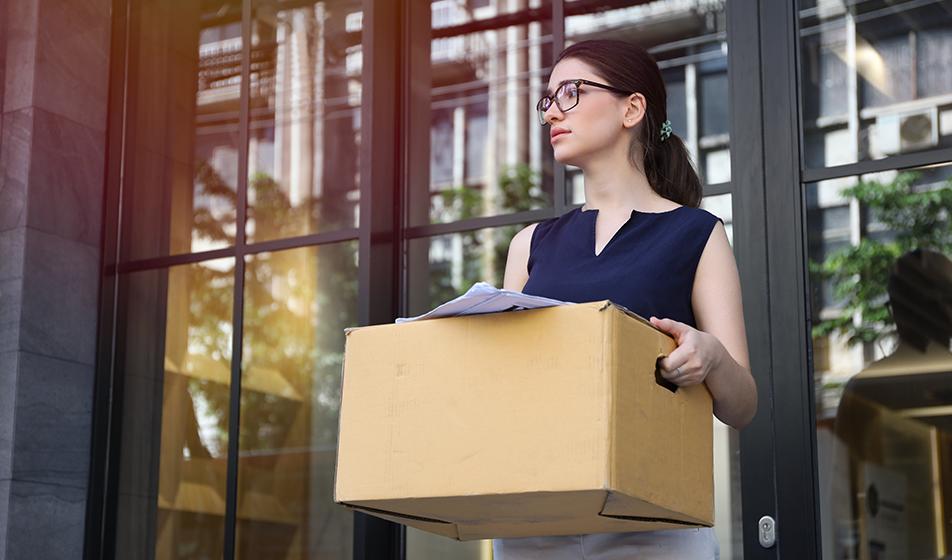 A woman stands outside an office building, holding a box containing desk supplies.