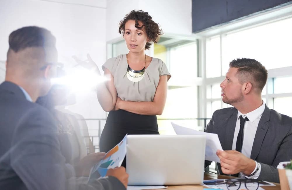 Three colleagues are working around a desk together. One colleague is stood up and talking to her other colleagues who have a laptop and paperwork in front of them.