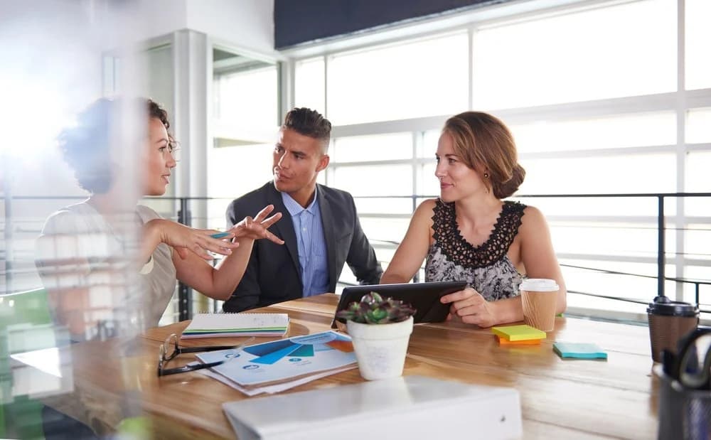 A team of three are having a business meeting. They are all sat at a wooden desk with a laptop and a notebook in front of them. 