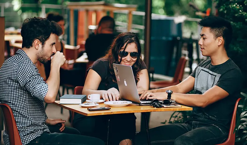 A group of colleagues are sat chatting at a wooden table outside of a coffee shop. They have a laptop, a book and their drinks in front of them.
