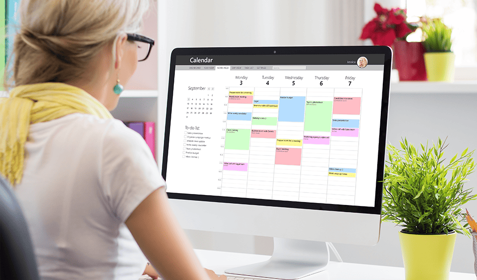 A woman sits at her desk looking at her work calendar on her computer screen. The back of her head can be seen and the screen is in full view, showing different-coloured meetings or blocks of time. 